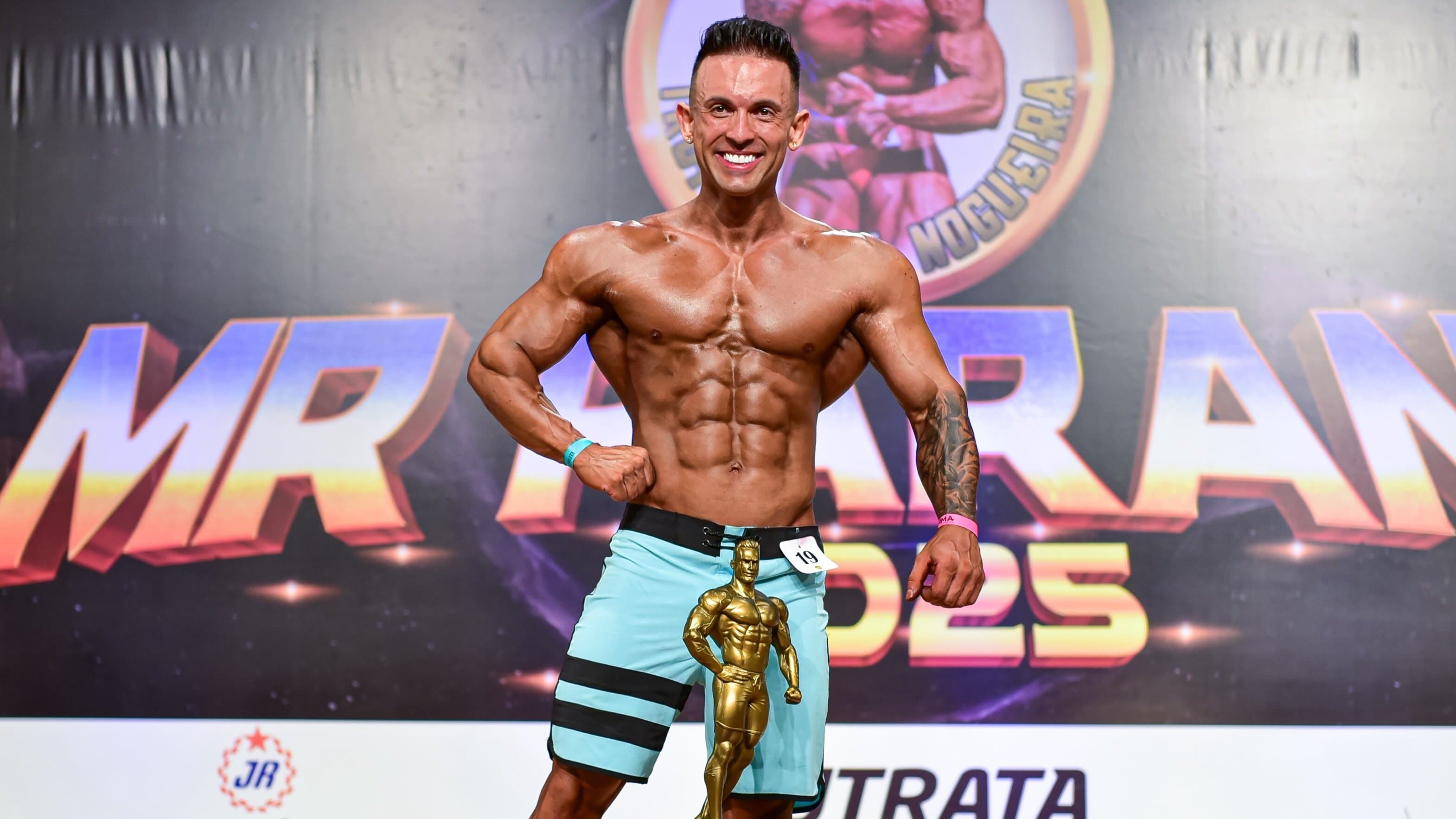 Bruno Rosa no palco de competição, campeão mundial de fisiculturismo, segurando o troféu de 1º lugar